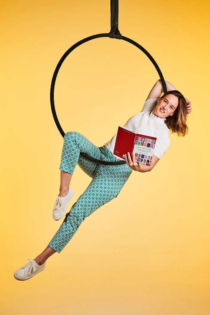 Dr Fleur van Rens in a lyra holding her book 'Circus psychology'. The background is bright yellow, her pants are teal, her top is white. 